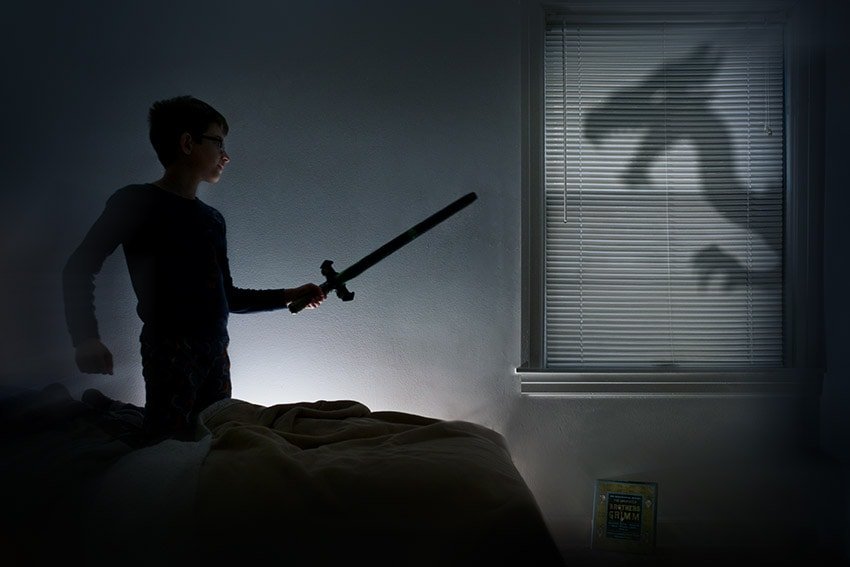 Photo of a boy with a toy sword facing the shadow of a dragon outside his window.