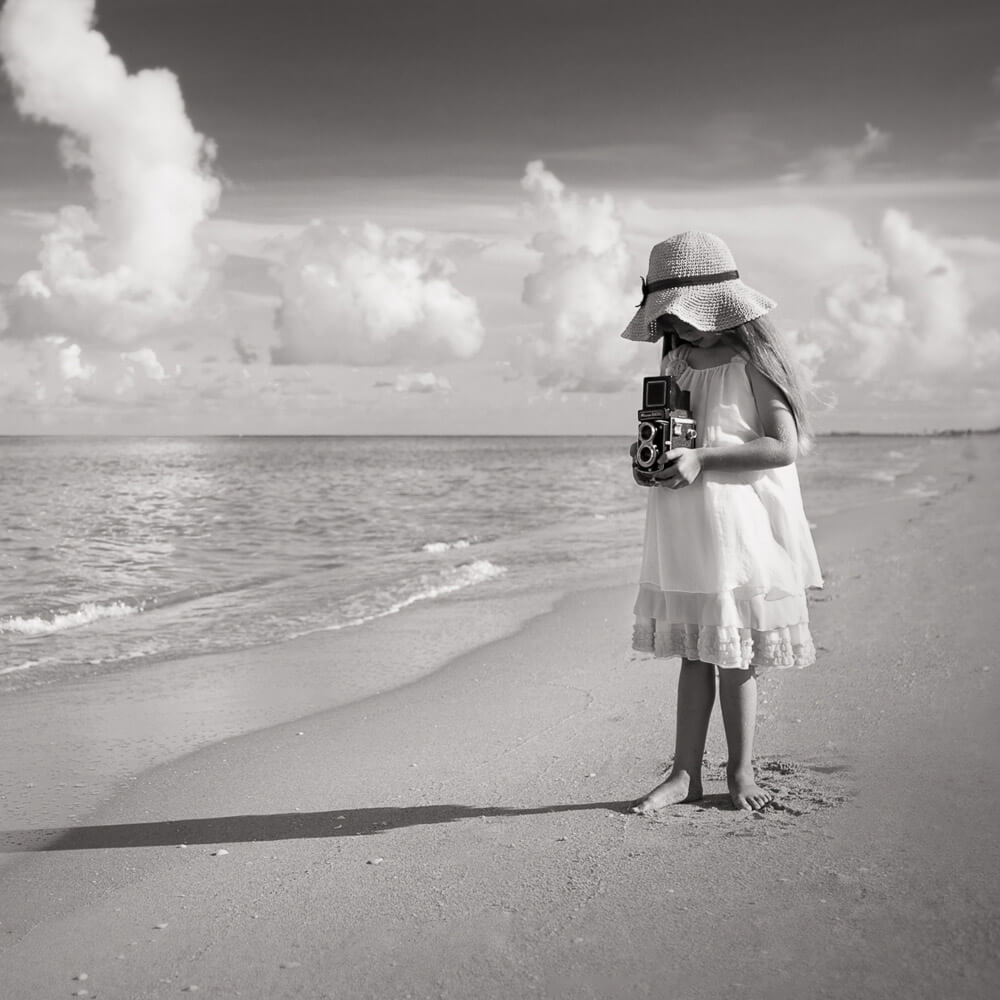 Black and white photo of a little in a dress and hat standing on the beach looking down into an antique camera | Quill & Glass blog